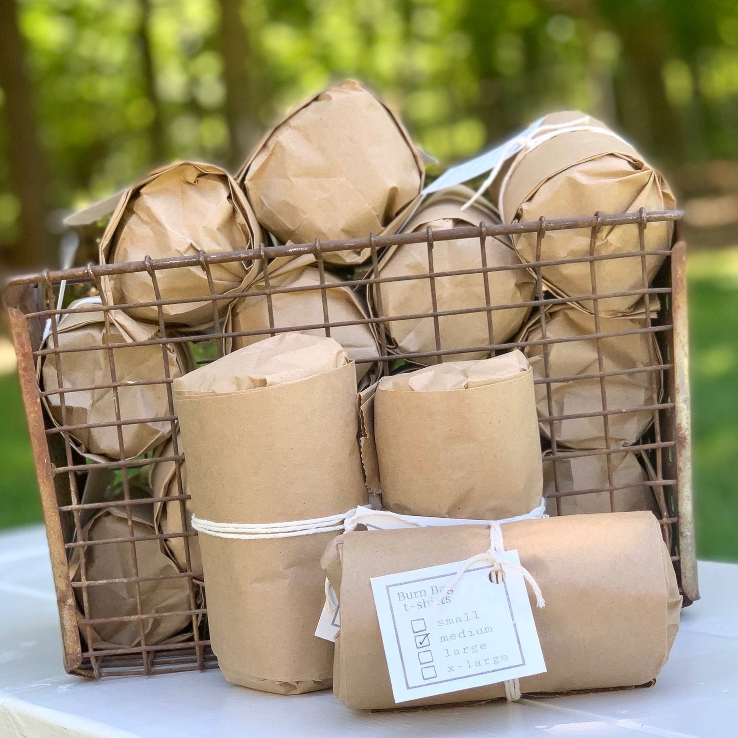 Stack of rolled charcoal gray “Burn Bags USA” T-shirts arranged in a wire basket with trees in the background.