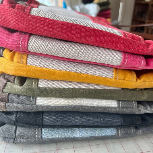 Stack of folded Classic Burn Bags in assorted colors—red, mustard yellow, olive green, gray, and blue—showing the variety of waxed canvas and hose panel combinations.