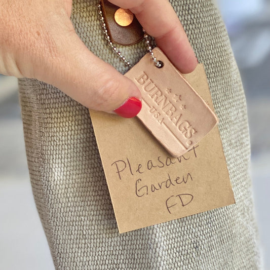 Person holding the Burn Bag Wine Carrier by its leather strap, showing the decommissioned fire hose body and attached leather tag.