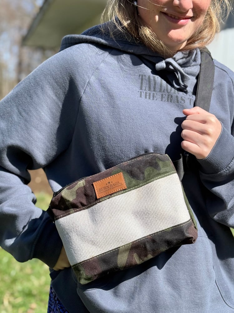 Person wearing the Special Edition Camo Fanny Pack crossbody and holding the strap, showing the reclaimed fire hose panel and BurnBags label.