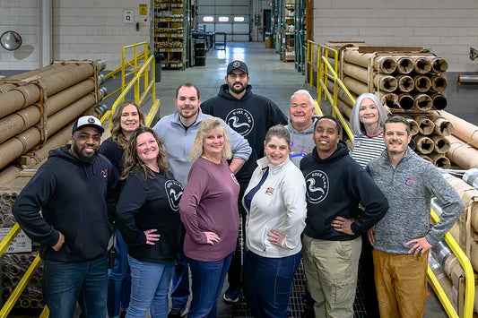 Team of eleven Eder Flag employees standing together in their manufacturing facility, surrounded by material used for flagpole production.