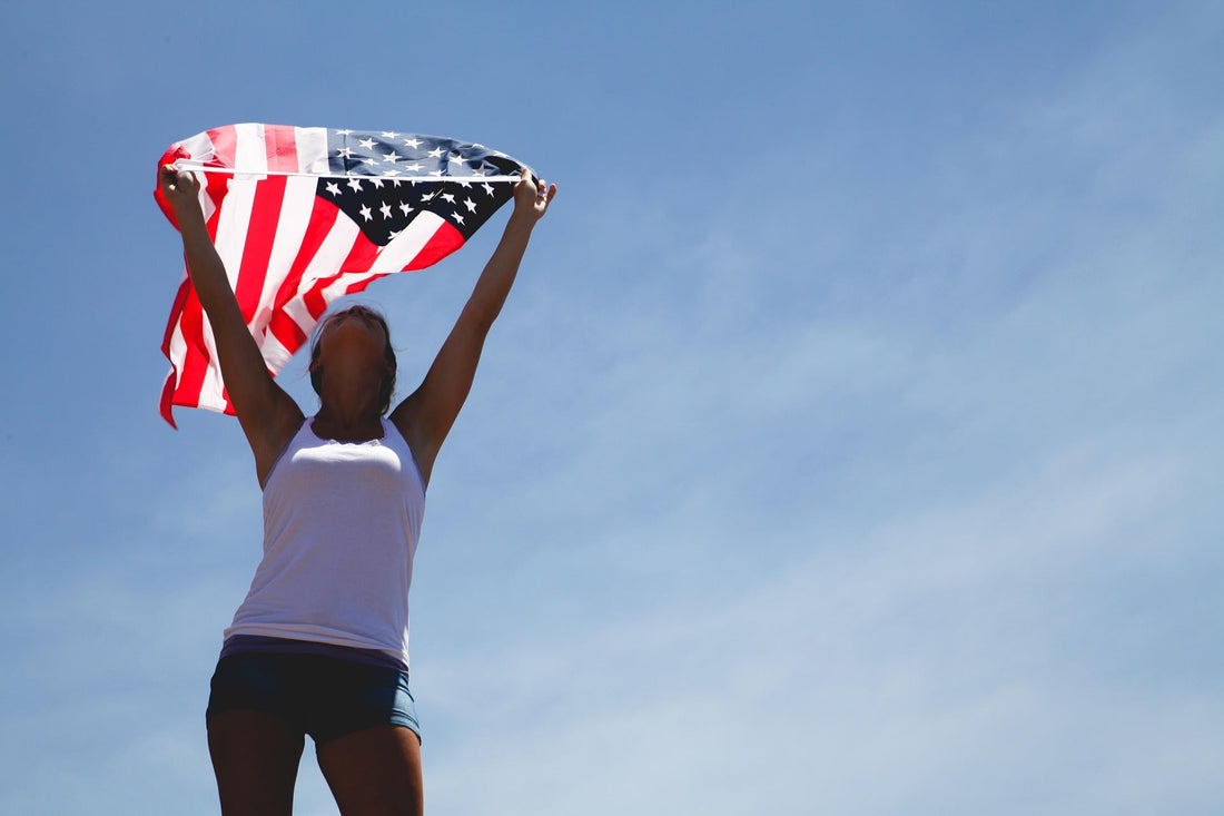 Woman standing outdoors under a clear blue sky, holding an American flag overhead as it waves in the sunlight, symbolizing patriotism, freedom, and national pride.