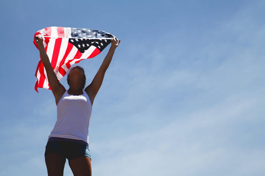 Woman standing outdoors under a clear blue sky, holding an American flag overhead as it waves in the sunlight, symbolizing patriotism, freedom, and national pride.