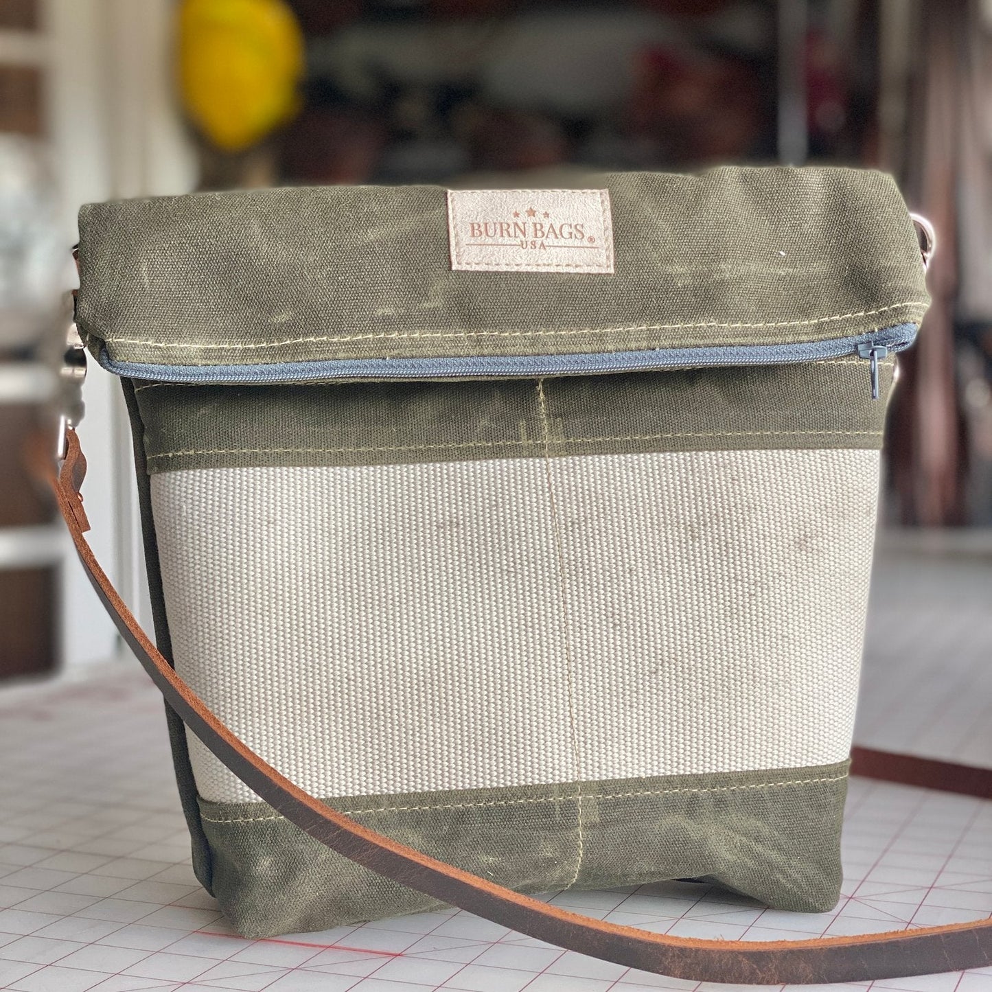 Olive green Crossbody Burn Bag shown standing on a sewing worktable, featuring a fold-over top, light hose panel on the front, and brown leather crossbody strap.
