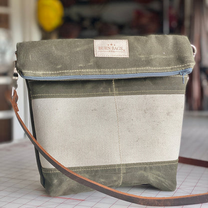 Olive green Crossbody Burn Bag shown standing on a sewing worktable, featuring a fold-over top, light hose panel on the front, and brown leather crossbody strap.