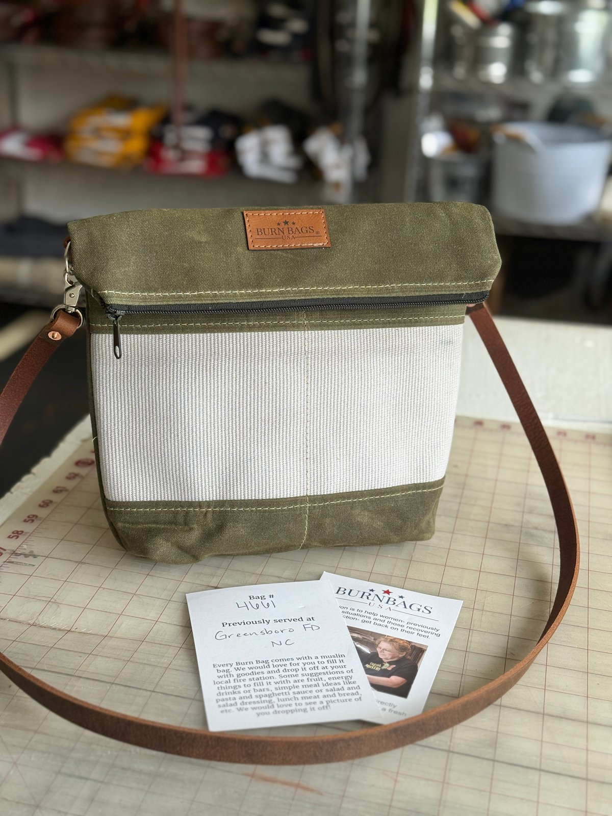 Olive green Crossbody Burn Bag shown upright on a sewing table with leather strap extended outward, accompanied by an authenticity card and story card placed in front of the bag.