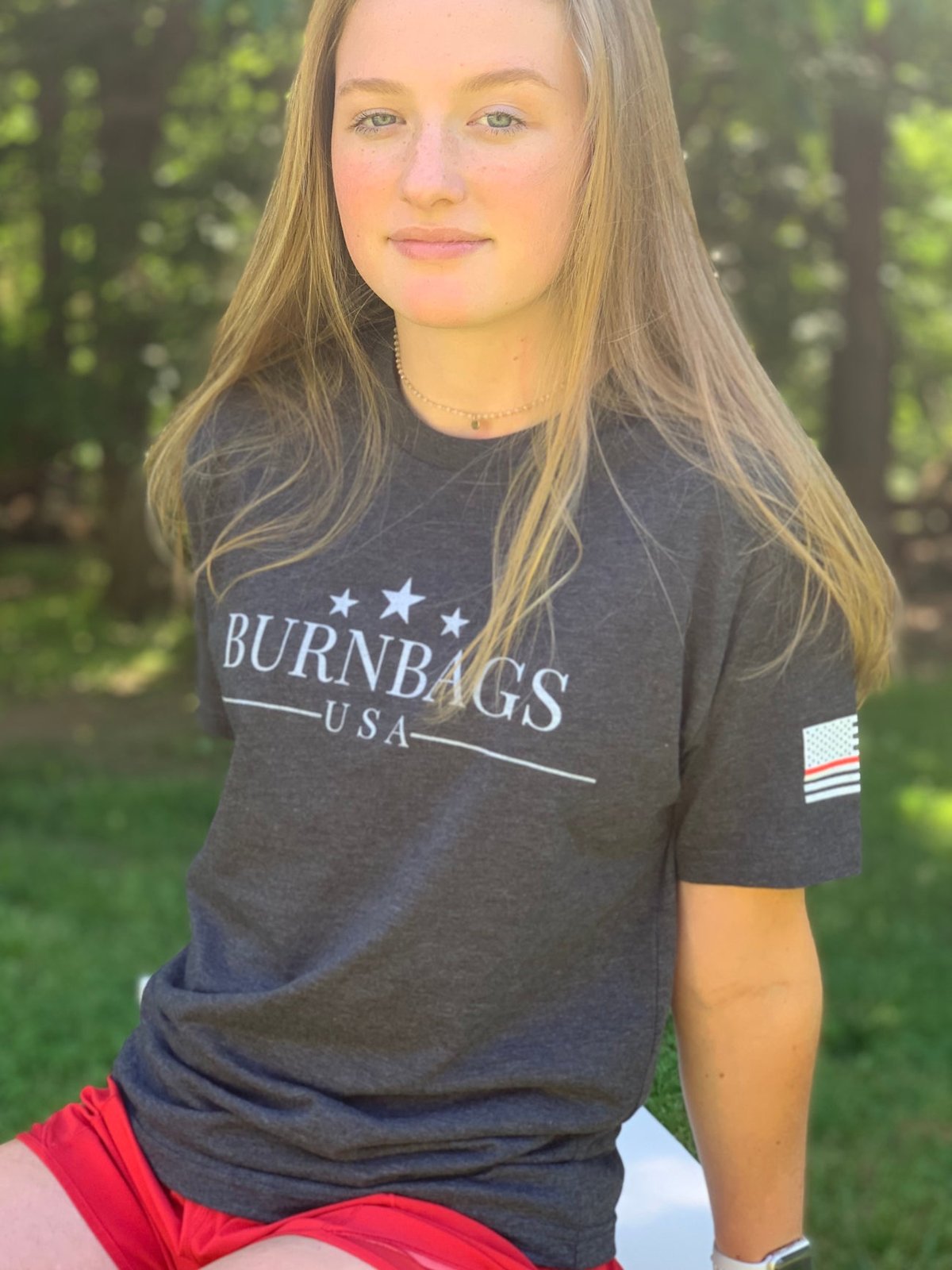 Person sitting outdoors wearing the charcoal gray “Burn Bags USA” T-shirt, showing the full front design and relaxed fit.