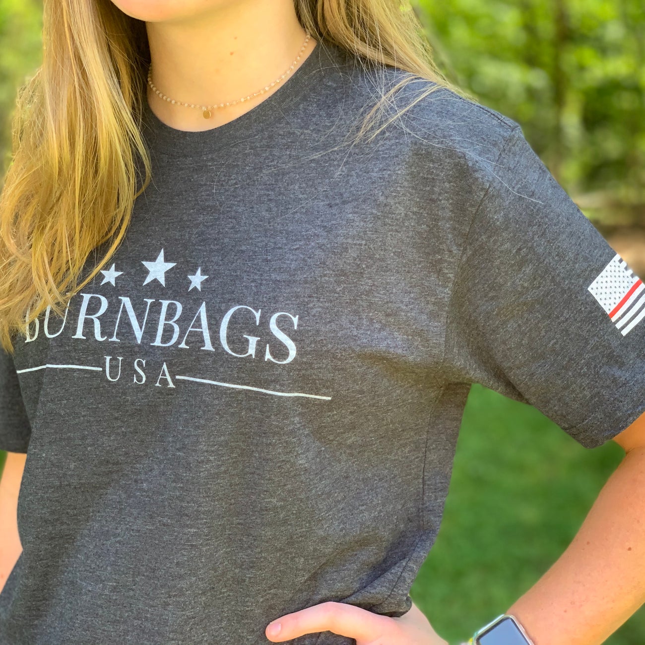 Person wearing a charcoal gray “Burn Bags USA” T-shirt outdoors, showing the front logo and American flag patch on the sleeve.