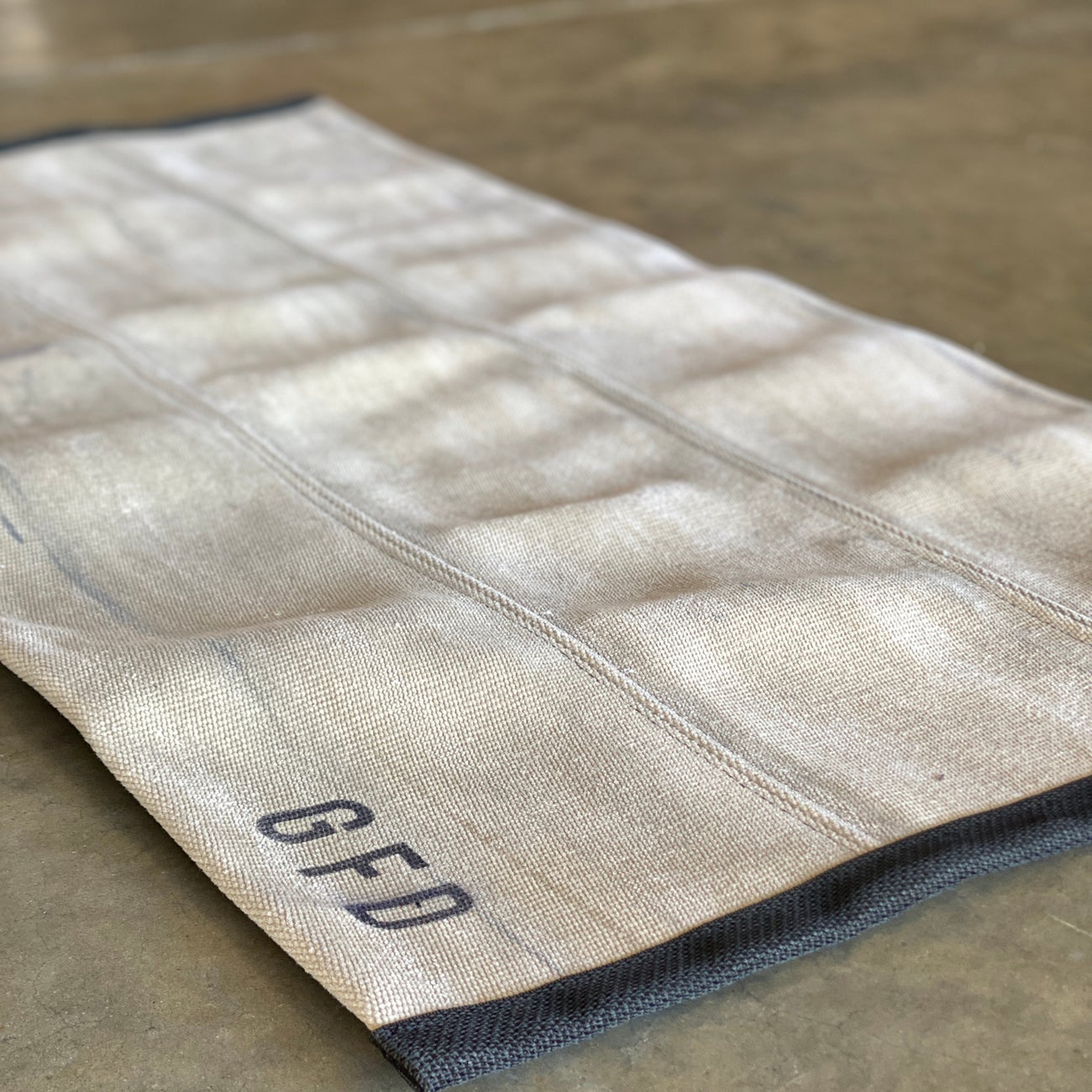 Khaki fire hose rug laid flat on a concrete surface showing textured hose panels and stamped with the department they are from on one end.