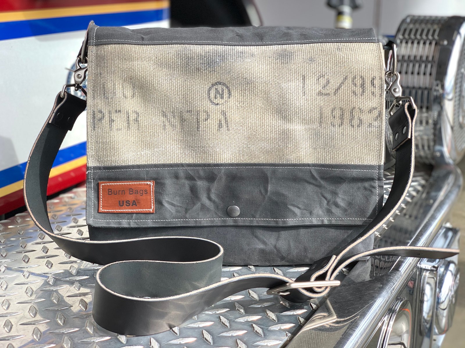 Charcoal Gray Messenger Burn Bag made from decommissioned fire hose and waxed canvas, displayed on the metal step of a fire truck with the flap closed and leather strap visible.