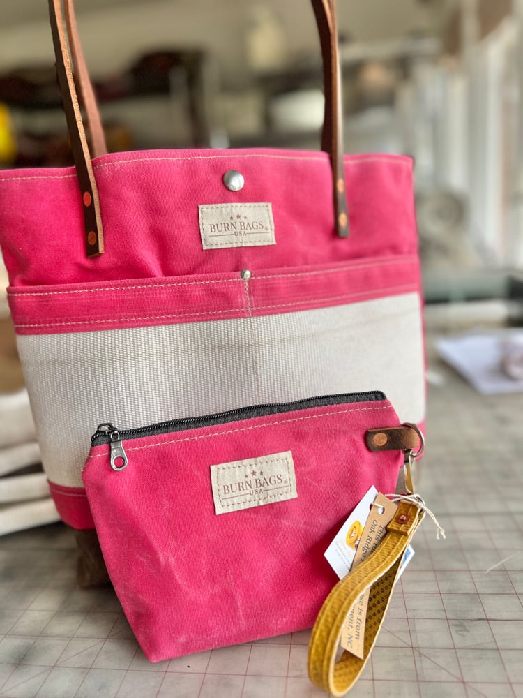 Peony Pink Classic Burn Bag displayed behind a matching pink zippered pouch with a wrist strap, both made from waxed canvas and decommissioned fire hose, shown on a worktable.